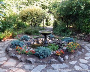 bird bath surrounded by flowers and grass and a path of stone around it